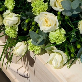 Sargschmuck mit weißen Rosen und grünen Nelken auf hellem Holzsarg bei Beerdigung.